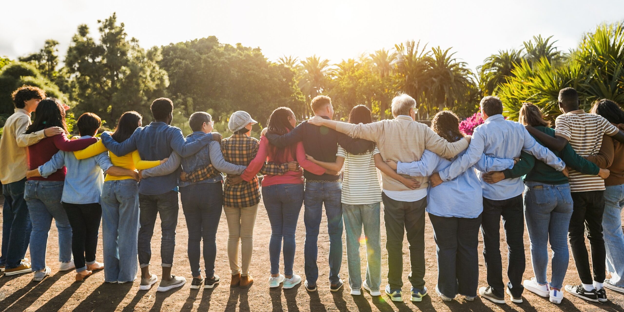A diverse group of people standing outdoors with arms around each other’s shoulders, facing the sunlight in a park setting.
